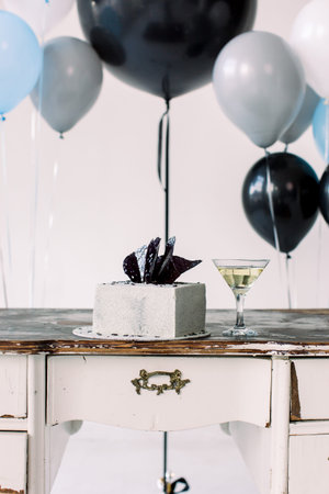 Close up photo of birthday cake and martini cocktail on the wooden table. Big colorful air balloons...