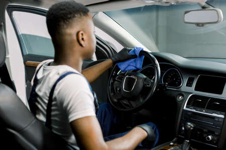Back view of young African man worker at car detailing service, wiping car steering wheel with microfiber cloth. Cleaning car interior with sanitizer and microfiber against virus infection.の写真素材