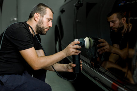 Car detailing and polishing concept. Professional Caucasian male car service worker, wearing black uniform, holding in hands orbital polisher, and polishing body of black luxury car.の写真素材