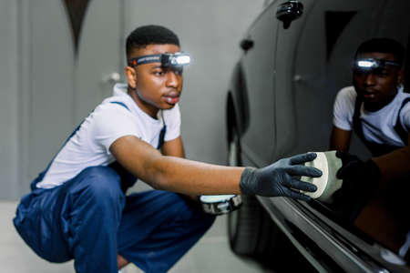 Car detailing, valeting concept. Close up of African man auto service worker, coating modern vehicle body with special professional wax, using the sponge. Selective focus on hand.の写真素材