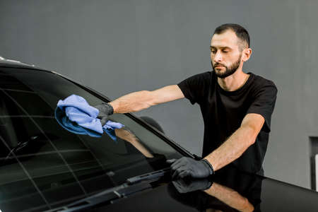 Car wash and cleaning at professional auto service station. Shot of handsome bearded young male worker in black t-shirt and gloves, cleaning car windscreen with blue microfiber cloth.の写真素材