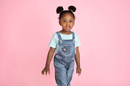 Cute little African preschool girl, wearing casual jeans overalls and t-shirt, posing to camera on isolated pink studio background. People, kids, studio portraits.の写真素材