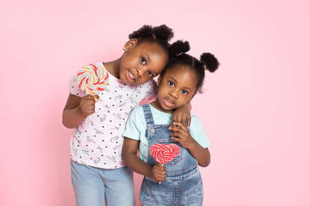 Two cute African girls sisters in casual summer outfits, posing with colorful lollipops hugging each other on pink studio background. Children and sweets.の写真素材