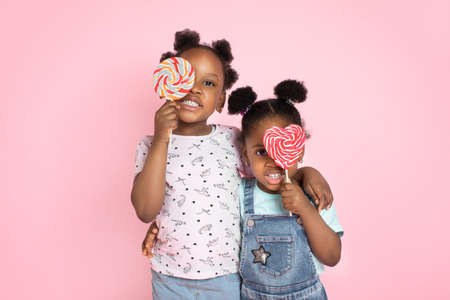 Close up horizontal shot of two cute little African girls covering her eyes with sweet colorful lollipops, smiling to camera on pink color background.の写真素材