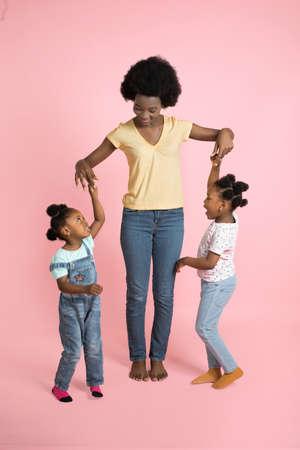 Portrait of happy family in colorful stylish outfits, charming young African woman mom and two cute little kids girls, having fun, playing and dancing together, on isolated pink background.の写真素材