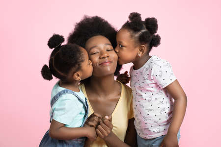 Motherhood and family concept. Lovely cute two little African girls kids in summer outfits, having fun and kissing their happy smiling young pretty mother, posing on isolated pink background.の写真素材