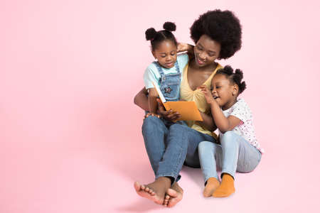 Family reading concept. Two adorable small african girls kids in colorful clothes, listening their pretty young mother reading to them stories from a book. Isolated on pink background, copy space.の写真素材