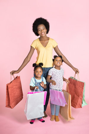 Full length portrait of pretty excited young African mother and her two little daughters, standing with many colorful shopping bags isolated over pink background. Family shopping.の写真素材