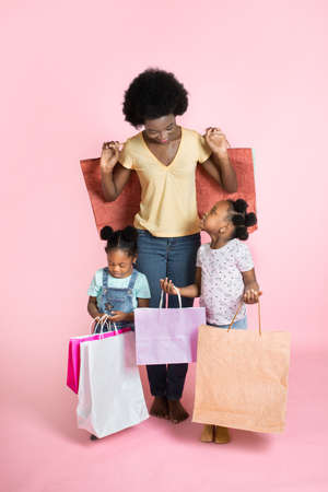 Beautiful young African mother and two litlle cute daughters in casual outfits, standing satisfied with shopping bags in hands, making purchases at the mall isolated on pink.の写真素材