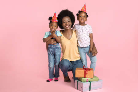 Portrait of young happy African mommy, hugging her two cute little kids daughters in party hats, celebrating birthday holiday party on pastel pink background with copy space.の写真素材