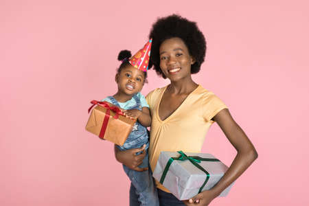 Close up of young pleasant African mother posing with her adorable cute little daughter in birthday hat, holding present boxes and smiling to camera over isolated pink background.の写真素材