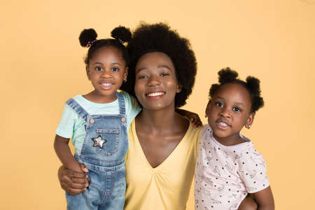 Family day concept. Close up studio portrait of happy young dark skinned mommy with her cute little two daughters, smiling, hugging and having fun together, on isolated yellow background.の写真素材