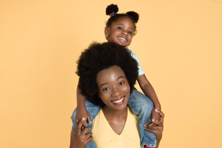 Fun with mom, happy childhood. Pretty African American mother piggybacking her daughter and smiling at camera. Isolated yellow background with copy spaceの写真素材