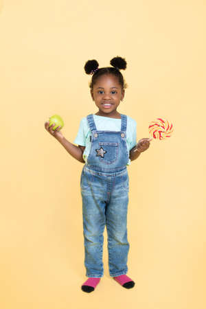 Little adorable funny black kid girl, wearing jeans overalls, chooses between a lollipop and a green apple, posing to camera with smile on yellow backgroundの写真素材