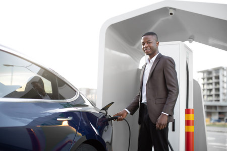 Handsome African American young business man plugging cable to electric car, posing to camera with...