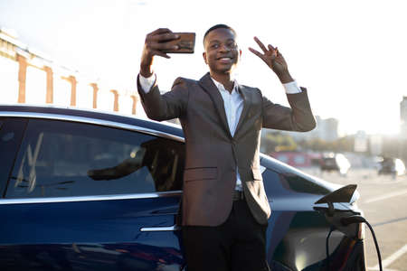 Smiling cheerful African business man, having video call or making selfie photo showing peace sign, standing near his electric car at city EV charging stationの写真素材