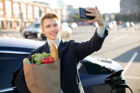 Portrait of young handsome european man, making selfie photo on his smartphone while leaning on his electric car, charging the battery at city power station after buying frsh products in trade mall.の写真素材