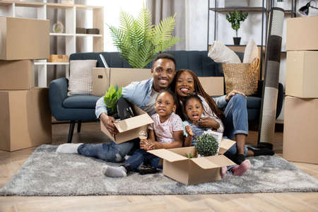 Cheerful african parents and cute two little daughters, sitting together on floor with boxes around. New life in new apartment. Concept of moving and property.の写真素材