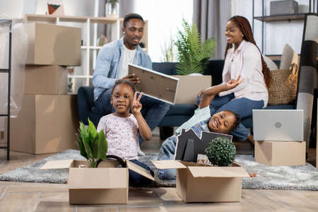 Young african couple sitting on couch and unpacking moving boxes while their pretty little daughters playing together on floor. Concept of relocation and mortgage.の写真素材