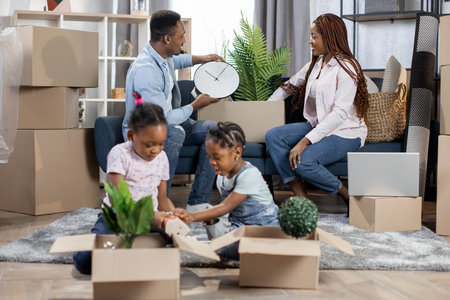 Afro american man and woman sitting together on sofa with boxes around. Two cute kids playing...