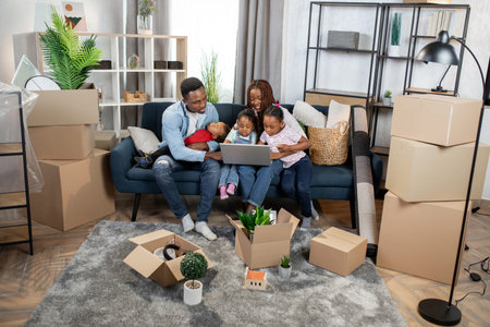 Young afro american parents with their three kids sitting together on couch and using modern laptop....
