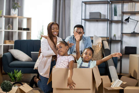 Two pretty kids sitting inside cardboard box and waving on camera while parents sitting on couch on background. New apartment for big family. Moving day.の写真素材