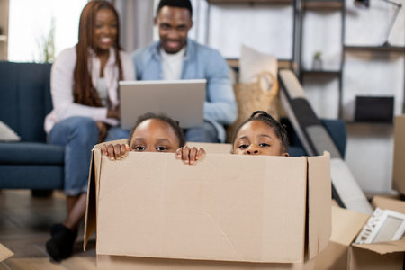 Two little african girls sisters looking at camera from cardboard box while their parents sitting on...
