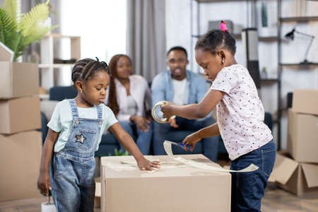 Two cute african kids packing boxes for moving to new house. Blur background of young parents sitting on couch and taking break. Relocation day.の写真素材