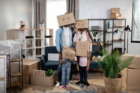 Young parents with their two kids standing together at new bright flat with cardboard boxes on...