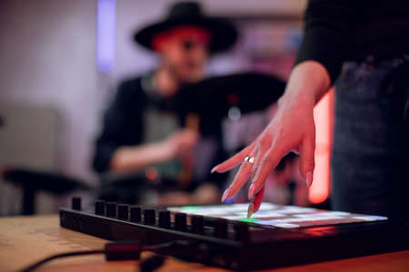 Close up of female hand touching mixing board for creating hit song at studio. Blur background of male artist playing on electronic drums. Music record.の写真素材