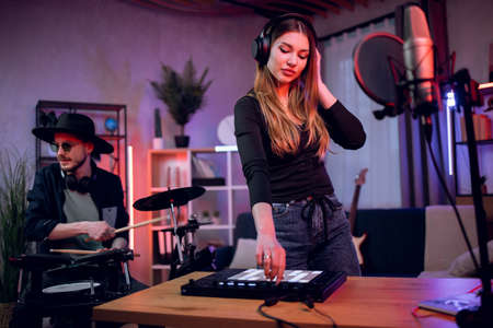 Adorable young woman in headphones working on mixing board while stylish man in hat and sunglasses playing electronic drums. Creative process in recording studio.の写真素材