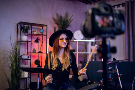 Stylish woman recording music video blog while playing electronic drums at home. Female musician in hat and glasses using digital camera indoors.の写真素材