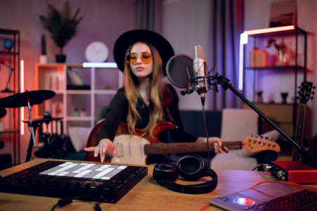 Attractive woman playing guitar and mixing sound on dj controller while sitting at modern studio. Female guitarist in stylish hat and glasses recording new song.の写真素材