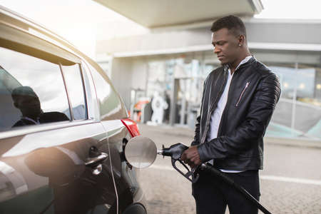 Portrait of young handsome bearded African man, wearing stylish casual clothes, refueling his luxury car at the gas station outdoors. Refueling of the car concept.の写真素材