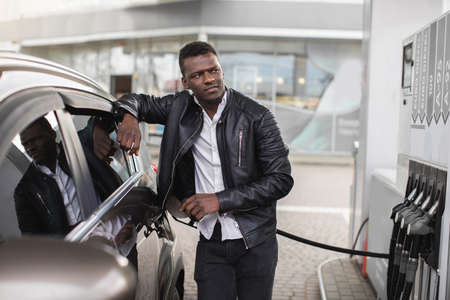 Front view of happy African man, leaning on his luxury modern car and looking away, while his car is refueling with gasoline at gas station.の写真素材