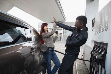Couple at gas station. Happy joyful multiracial couple of friends or colleagues, having fun while refueling luxury car at petrol station, giving high five each other and smiling.の写真素材