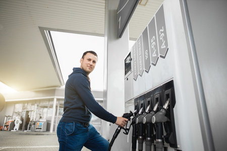 Young handsome man in casual clothes, refueling his car with gasoline at a gas station, taking...