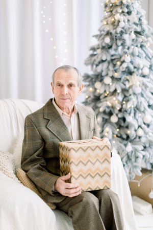 Handsome retired man in suit and shirt, holding present box, while sitting in white armchair in front of beautiful decorated Christmas tree at cozy indoor studio. Christmas presents concept.の写真素材