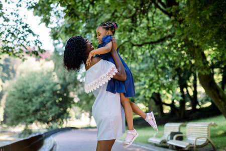 Young beautiful African woman mother in light blue dress playing with her little cute daughter in the park, having fun and lifting the baby up. Happy motherhood concept.の写真素材