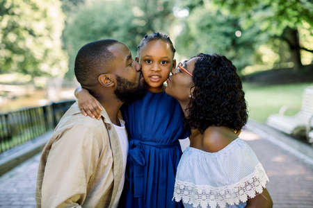Adorable summer portrait of young african family of three posing together outdoors in beautiful park. Lovely parents are kissing their little daughter in blue dress. Close up.の写真素材