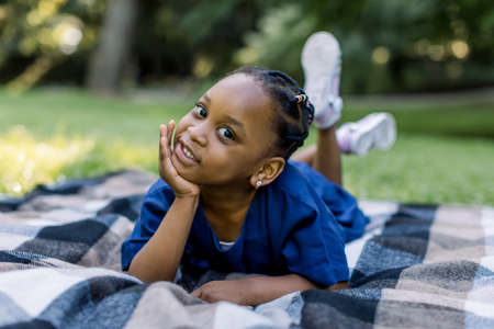 Outdoor portrait of a cute little African girl lying down on the checkered blanket in the park and smiling. Adorable child having fun on a walk.の写真素材
