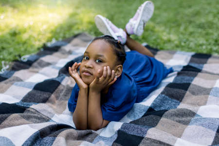 Happy smiling African girl child lying on blanket in summer park, leaning her face on her hands and looking at camera. Happy African child resting on green grass in park.の写真素材