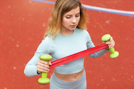 Young attractive professional woman fitness trainer, doing exercise with dumb bells and red rubber...