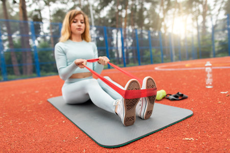 Attractive fitness woman has a workout outdoors in the red court, sitting on fit mat and using...
