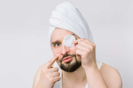 Funny face of handsome bearded man with a towel on his head posing to camera with cotton pad covering his one eye. Skin care and dermatology. Spa for men.の写真素材