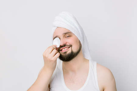 Smiling handsome guy with a white towel on his head cleaning face skin with cotton pad. Attractive man with a pad covering his eye looking at the camera, isolated on white. Close up photo, copy space.の写真素材