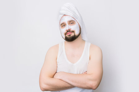 Handsome bearded man with white towel on his head and cosmetic mask on his face, stands with arms crossed on white background. Cosmetology procedures for men. Skin care for menの写真素材