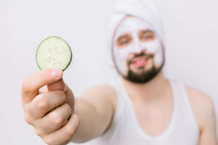 Blurred face of handsome bearded man in white towel having white mask on his face and posing with slice of cucumber, showing it to camera. Man with mask on face, white background, focus on cucumber.の写真素材