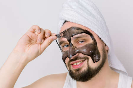 Handsome smiling bearded guy is removing purifying black mask from his face over white background. Close up portrait of man during spa treatment.の写真素材