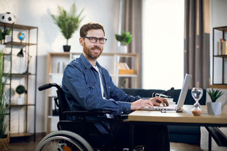 Side view of bearded young man sitting in wheelchair and typing on wireless laptop. Handsome disabled guy in eyewear working from home.の写真素材